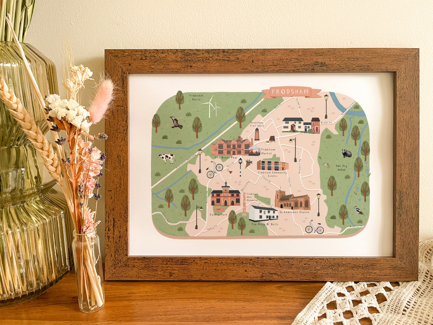 A wooden frame featuring a colourful illustrated map of Frodsham, styled on a wooden tabletop next to a champagne-coloured glass vase and dried flowers.