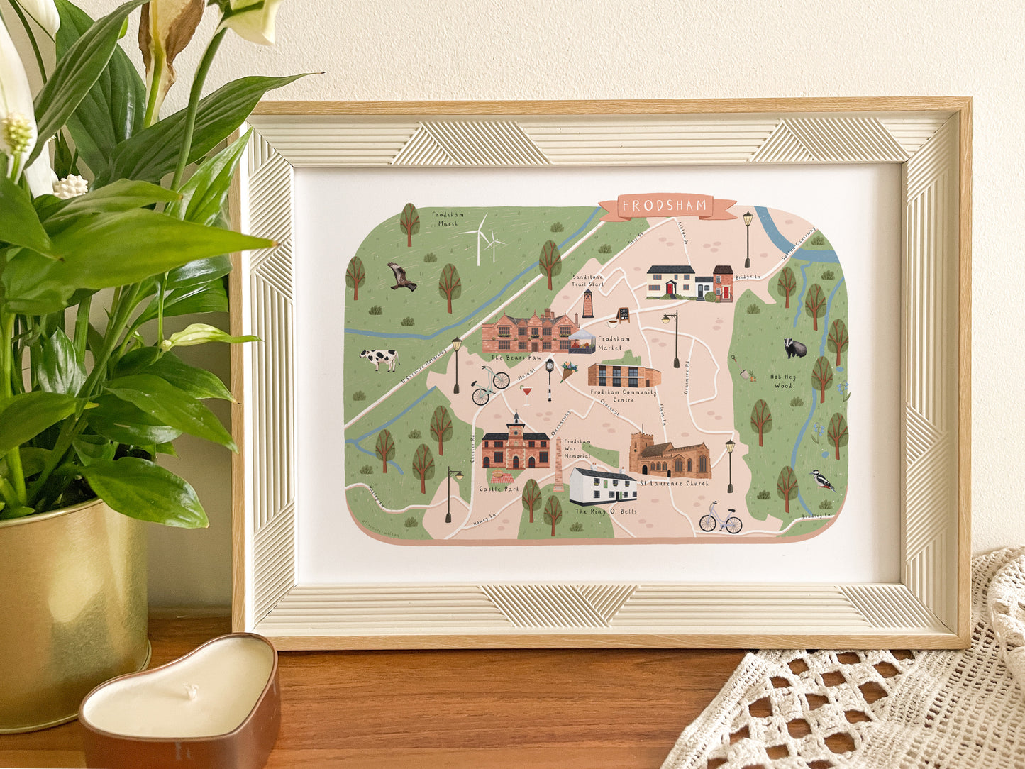 A framed art print featuring a whimsical illustrated map of Frodsham in Cheshire, styled on a wooden table next to a potted green plant.