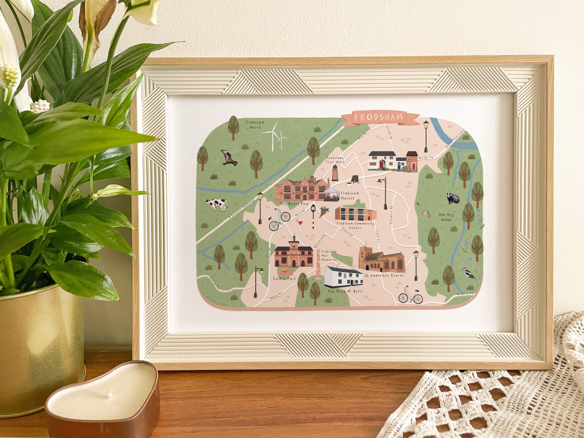 A framed art print featuring a whimsical illustrated map of Frodsham in Cheshire, styled on a wooden table next to a potted green plant.