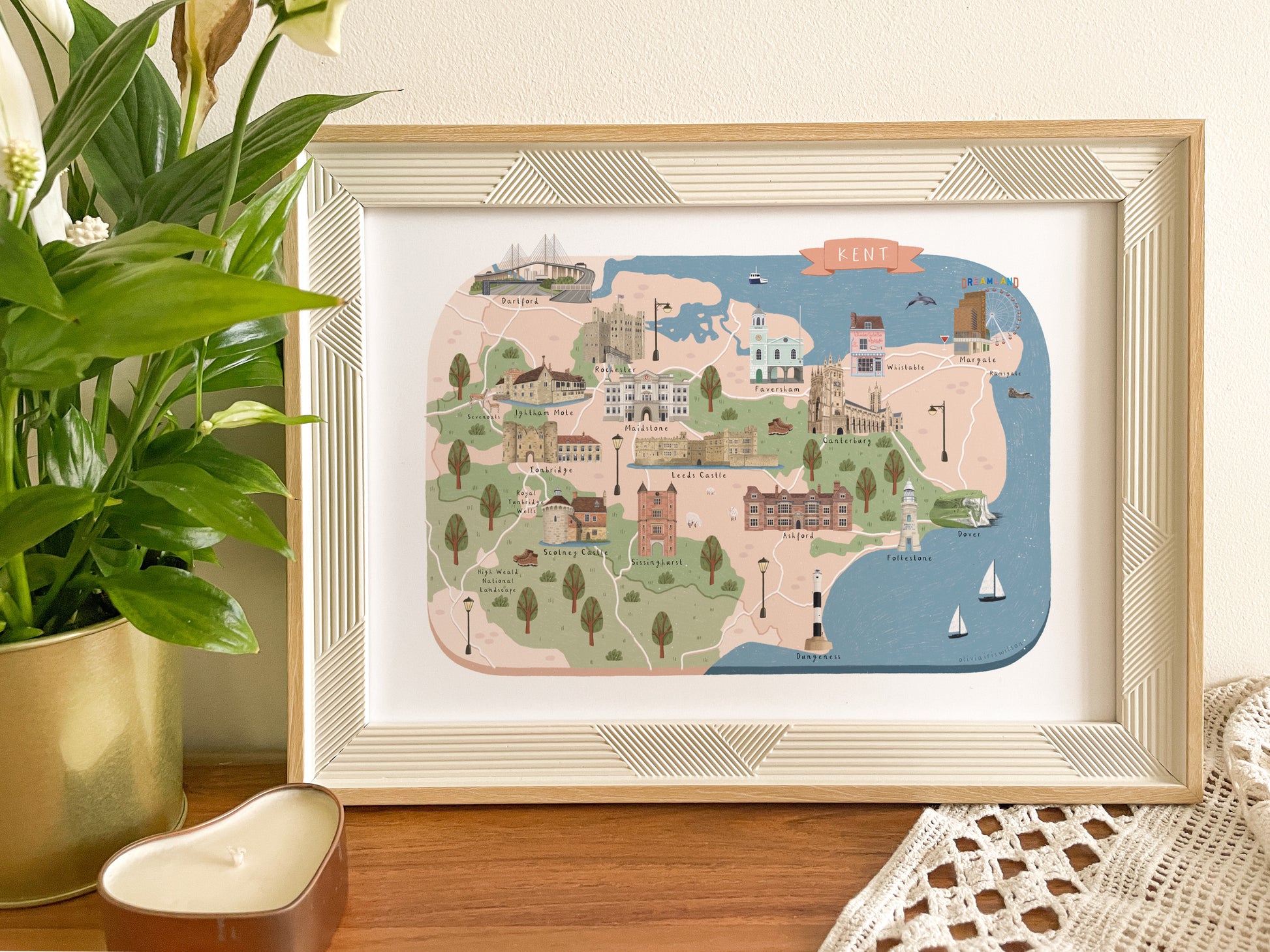 A framed illustration print of Kent detailing the coastline, displayed on a wooden surface next to a potted green plant. The print features a whimsical map of the towns and seaside locations of Kent. There are notable buildings, boats, and landmarks depicted in soft pastel colours, housed in a light wood frame with a white mat.