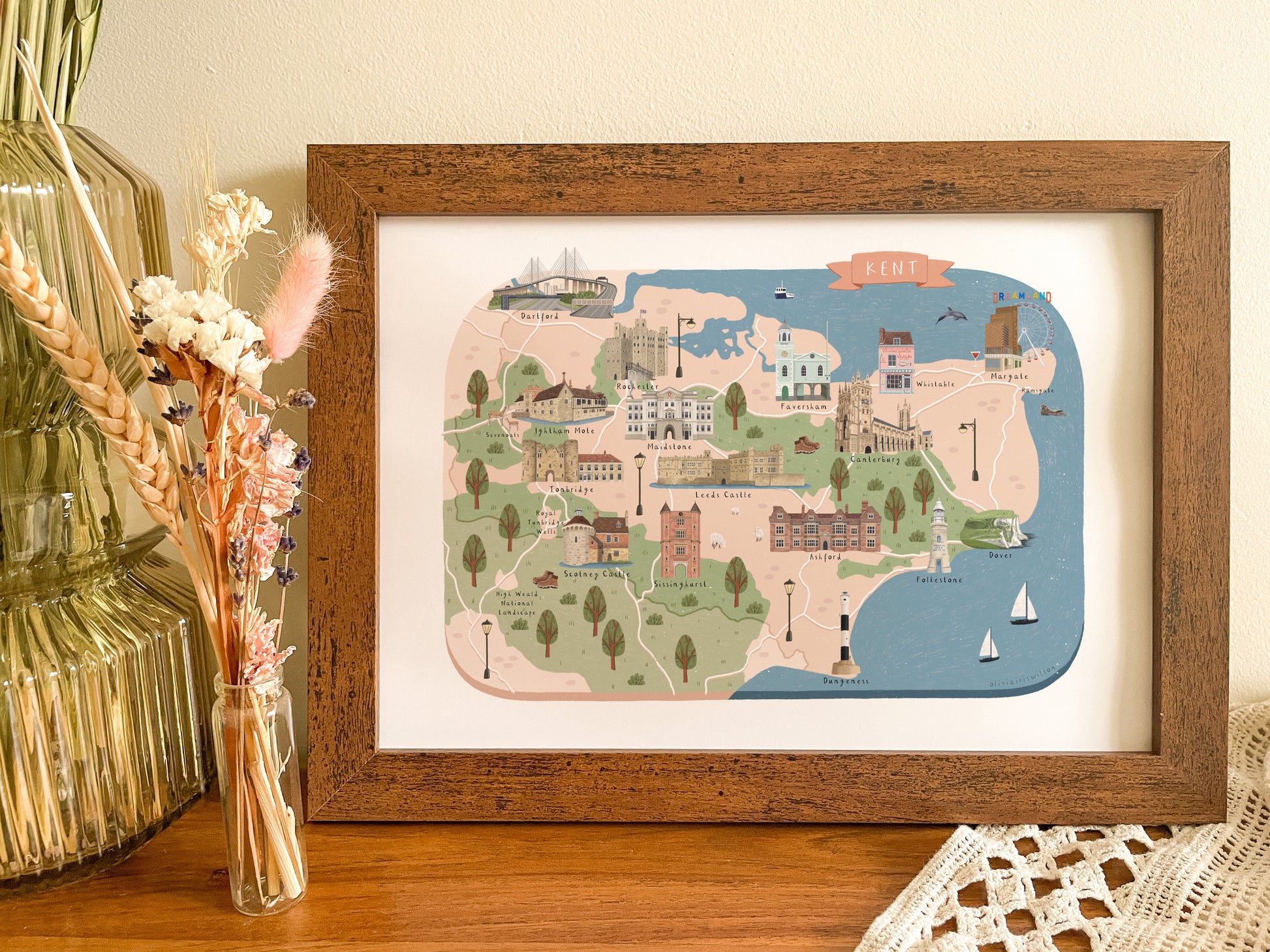 A framed art print featuring a hand-drawn illustrated map of Kent, England, styled on a wooden table next to a glass vase with dried flowers.
