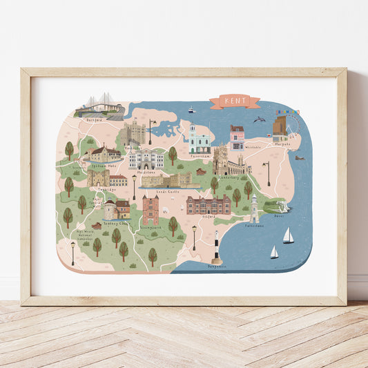 A colourful illustrated map of Kent, UK, featuring pastel-colored landmarks, buildings, trees, and the coastline, displayed in a light wood frame on a light wood floor against a white wall.