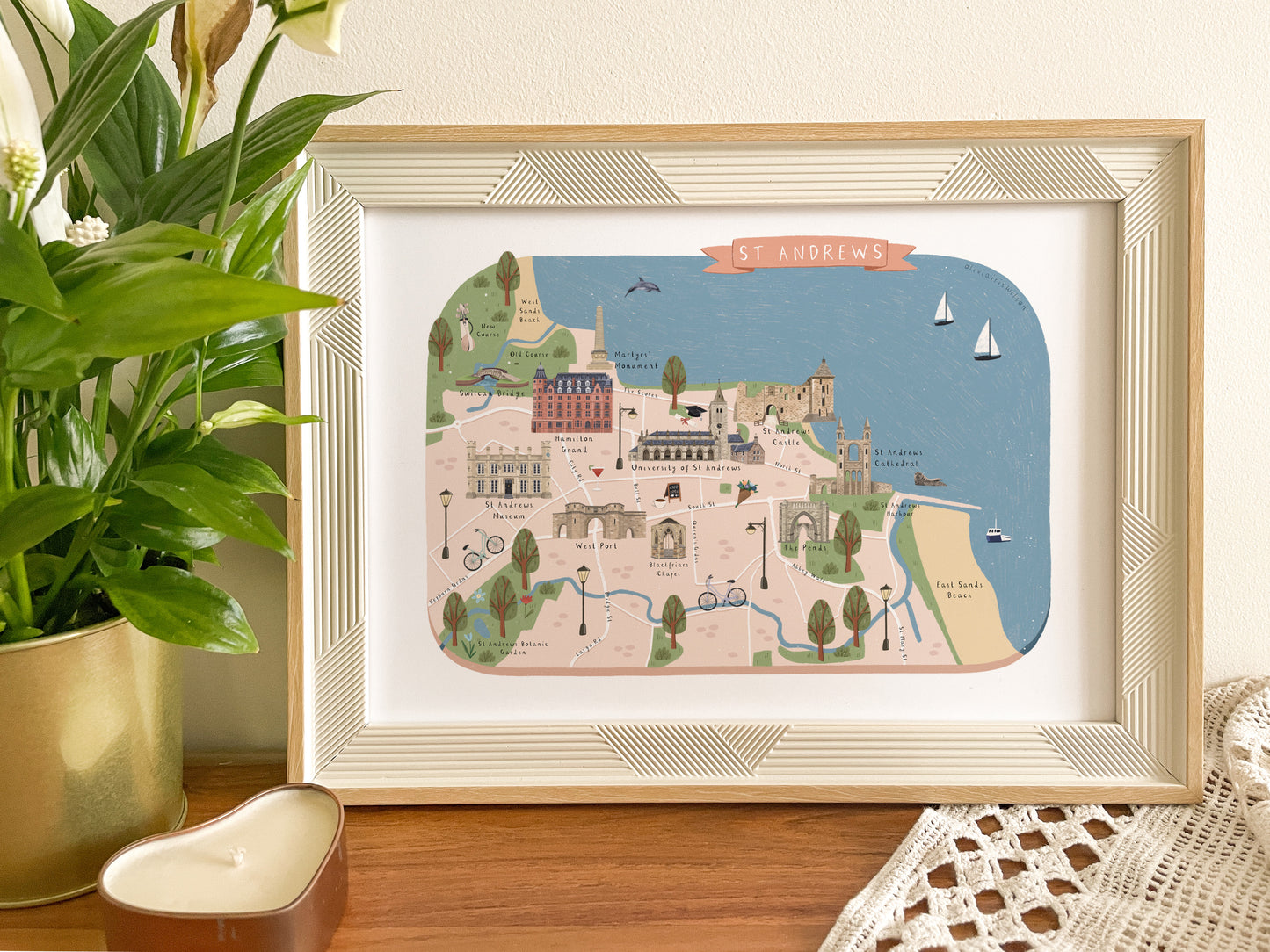 A framed art print depicting an illustrated map of St Andrews, styled on a wooden surface next to a potted green plant and a white crocheted textile.