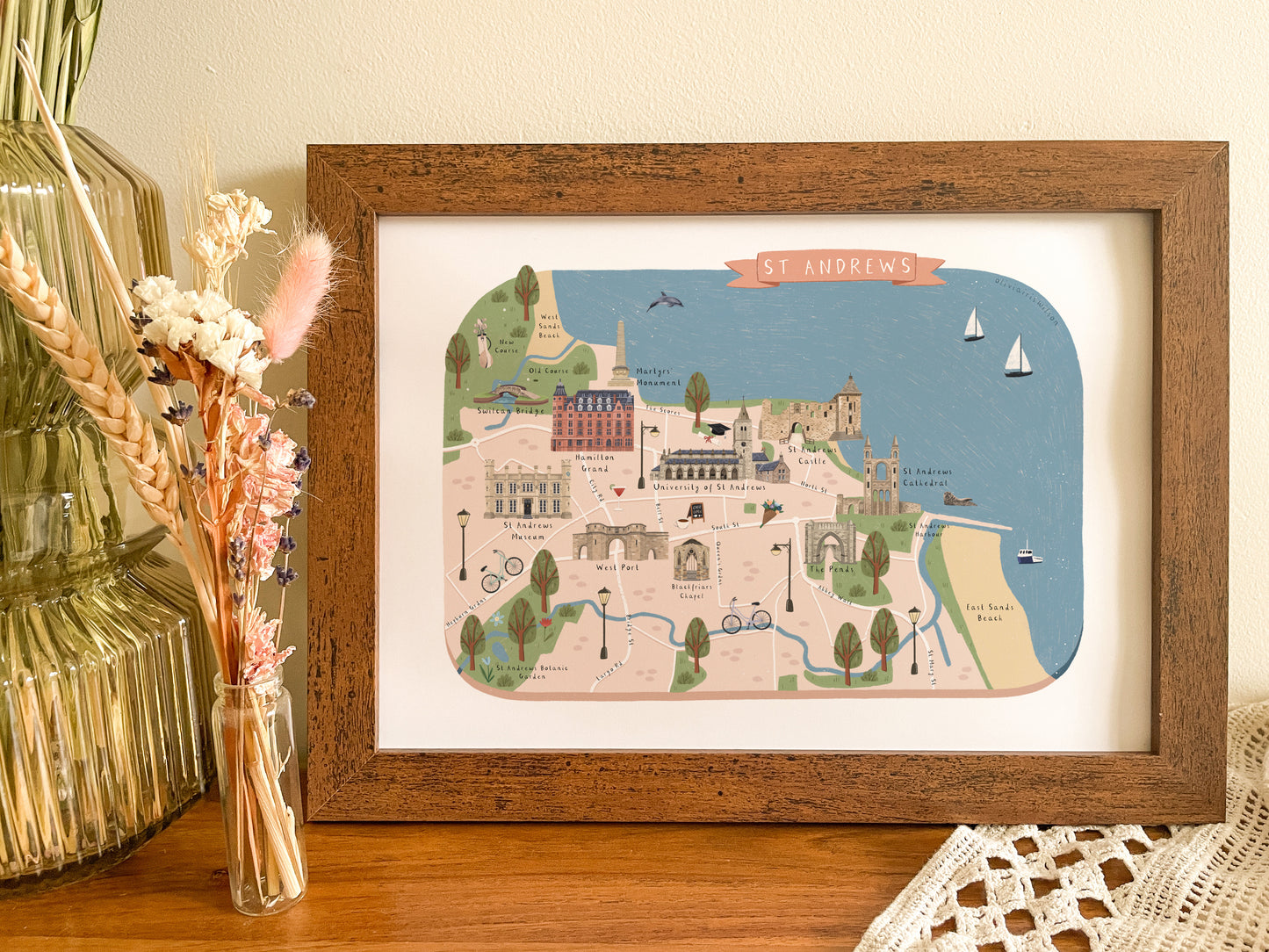 A framed art print featuring a hand-drawn illustrated map of St Andrews, styled on a wooden table next to a glass vase with dried flowers.