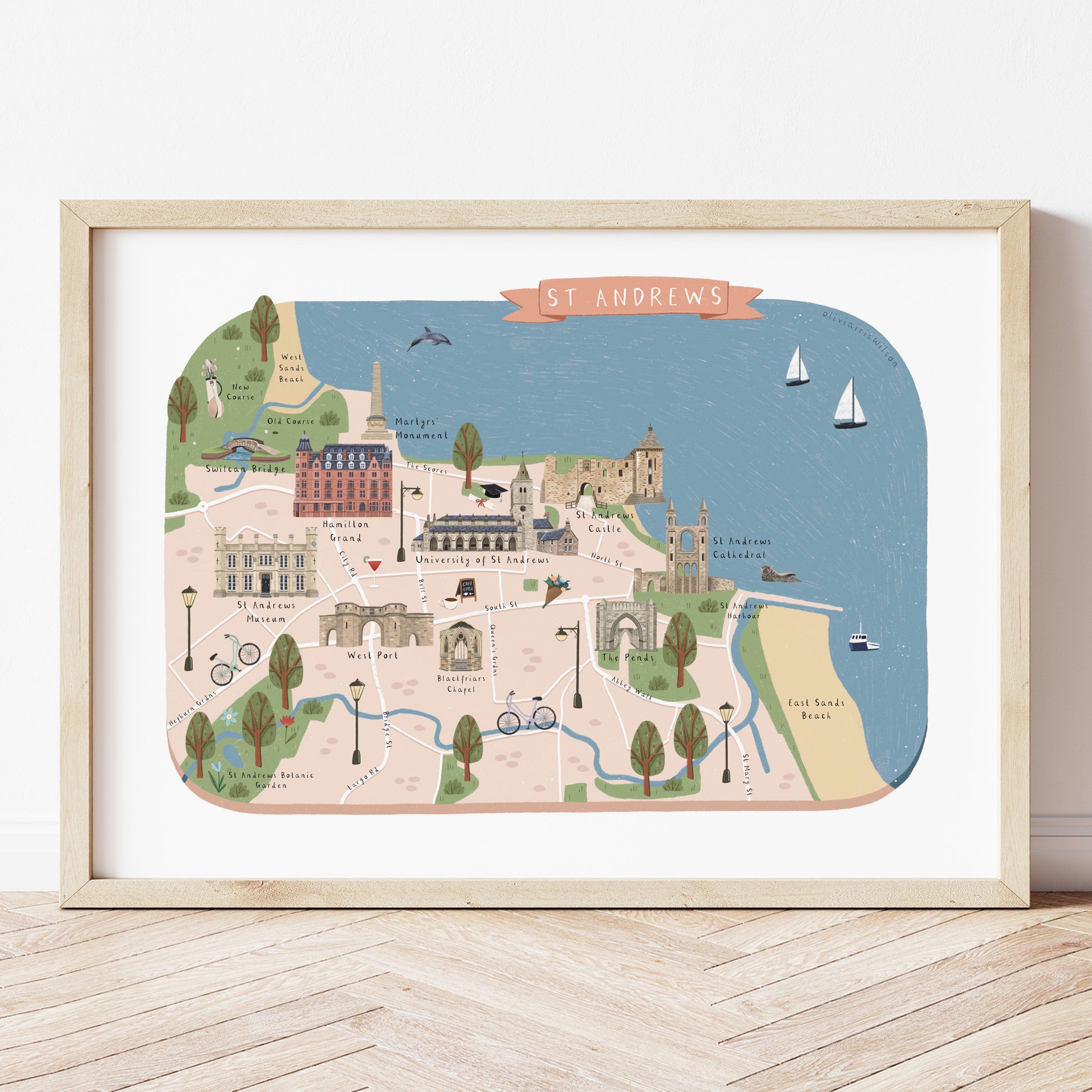 A framed St Andrews map art print with a light wood frame, leaning against a white wall on a light wood floor. The map features a colourful illustration of the cityscape, coastline, and landmarks in pastel colours.