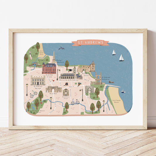 A framed St Andrews map art print with a light wood frame, leaning against a white wall on a light wood floor. The map features a colourful illustration of the cityscape, coastline, and landmarks in pastel colours.
