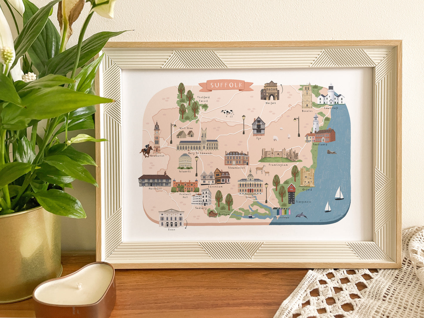 A framed illustrated map print of Suffolk, England, styled on a wooden surface next to a potted green plant and a white crocheted textile.