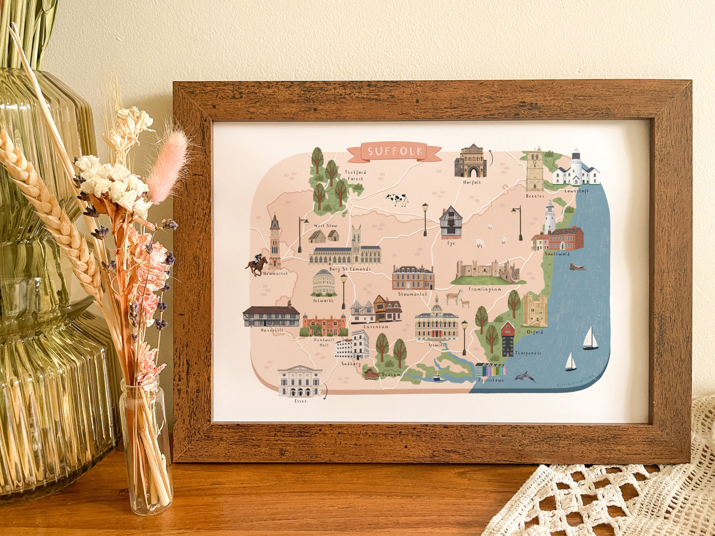 A framed illustrated map of Suffolk, England, displayed on a wooden surface next to dried flowers in a glass vase. The map features a warm beige background with various colorful illustrations of Suffolk landmarks, surrounded by a white mat and a rustic brown wooden frame.